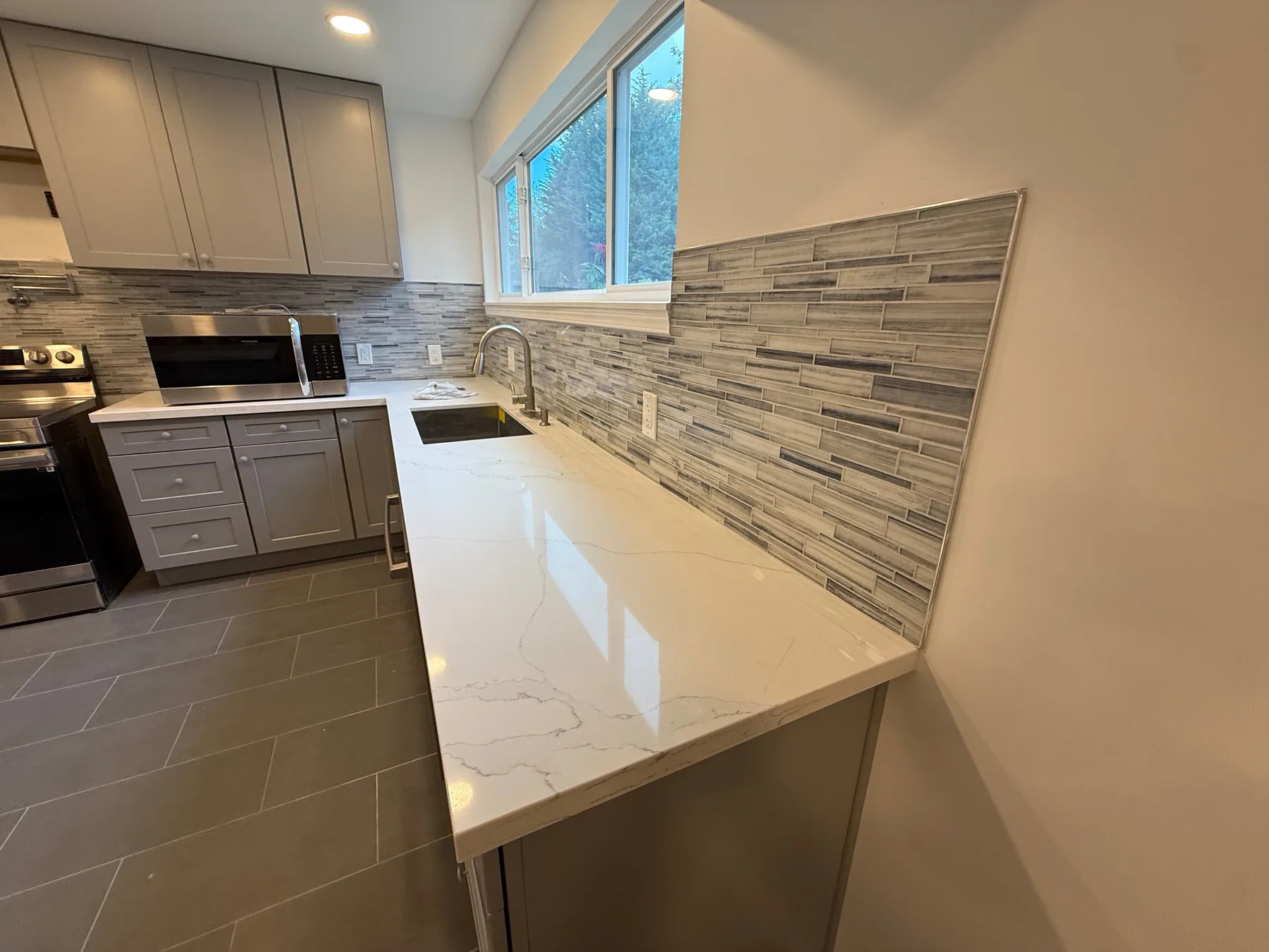 Gray kitchen cabinets, tiled backsplash, sink, and white countertop in a completed remodel